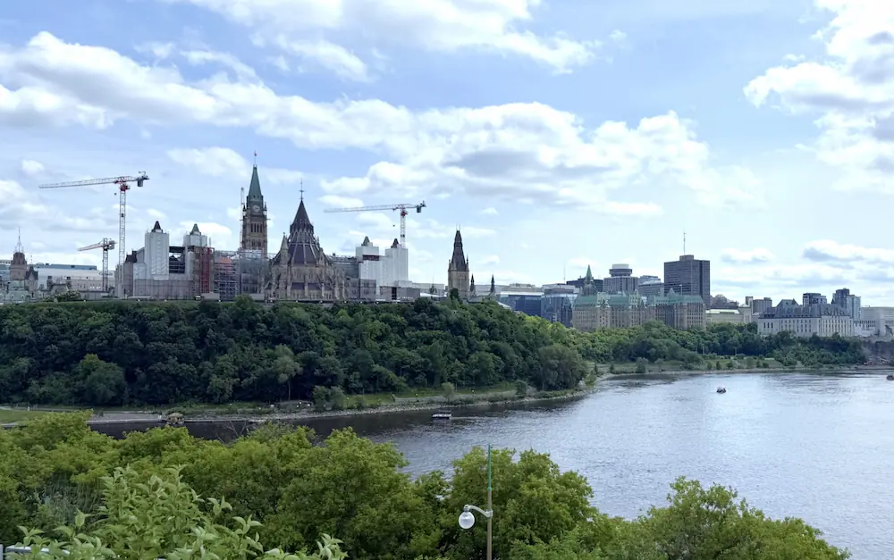 View of downtown Ottawa and Parliament Hill from Kiweki Point, with the Ottawa River in the foreground and construction cranes above the skyline.