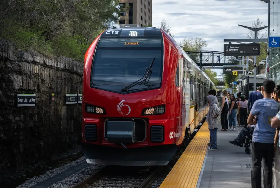 Passengers waiting on the Dow’s Lake O-Train platform as a red Stadler FLIRT train arrives on a sunny spring day.